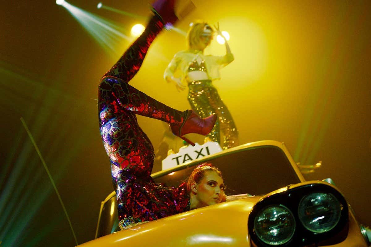 Dois artistas do espetáculo Mad Apple, do Cirque du Soleil em Las Vegas. Eles fazem poses dinâmicas em um táxi amarelo e ao redor dele, sob uma iluminação dramática no palco.
