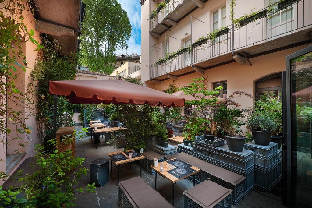 Terraço compartilhado do Maison Borella com vasos de plantas, mesas e bancos de madeira, um toldo vermelho, para representar os melhores hotéis em Milão