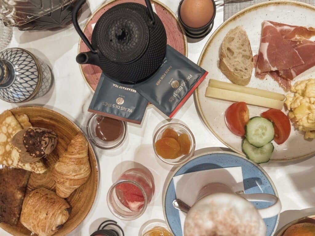 A imagem mostra uma mesa de café da manhã variado vista de cima na Maison Mère. Há uma chaleira preta com saquinhos de chá ao redor, um prato com presunto, queijo, fatias de pão, tomate e pepino, e ovos mexidos. Uma cesta contém croissants, pães de chocolate e panquecas. Há também um ovo cozido em um suporte, potes com damascos secos, geleia e iogurte, além de uma xícara de café com espuma de leite. A mesa está organizada de forma apetitosa e convidativa, com diversos itens para uma refeição completa.