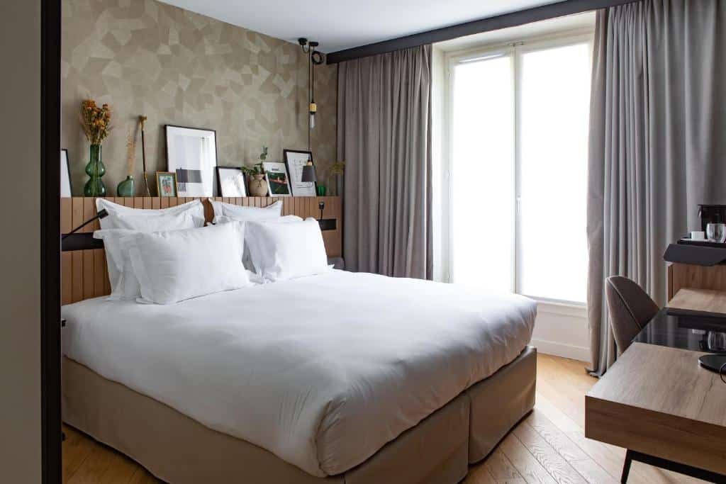 A imagem mostra um quarto moderno e aconchegante na Maison Mère, um dos hotéis com café da manhã em Paris. A cama é grande, com lençóis brancos impecáveis e várias almofadas. A parede atrás da cama tem um padrão geométrico discreto e está decorada com várias molduras e vasos com flores secas. Há uma janela alta com cortinas cinza, permitindo a entrada de luz natural. Ao lado da cama, há uma mesa de trabalho de madeira com uma cadeira e uma pequena área com cafeteira e copos. O piso é de madeira clara, complementando o ambiente acolhedor do quarto.