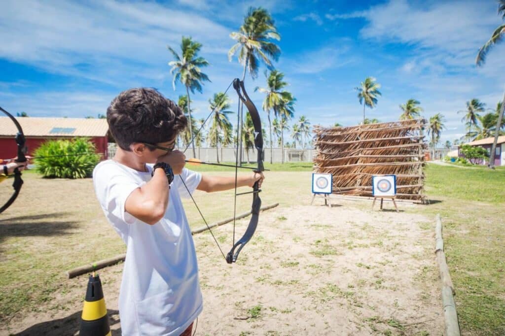 Uma pessoa praticando arco e flecha mira uma flecha em alvos em um dia ensolarado no Makai Resort All Inclusive Convention Aracaju. Palmeiras e estruturas são visíveis ao fundo.