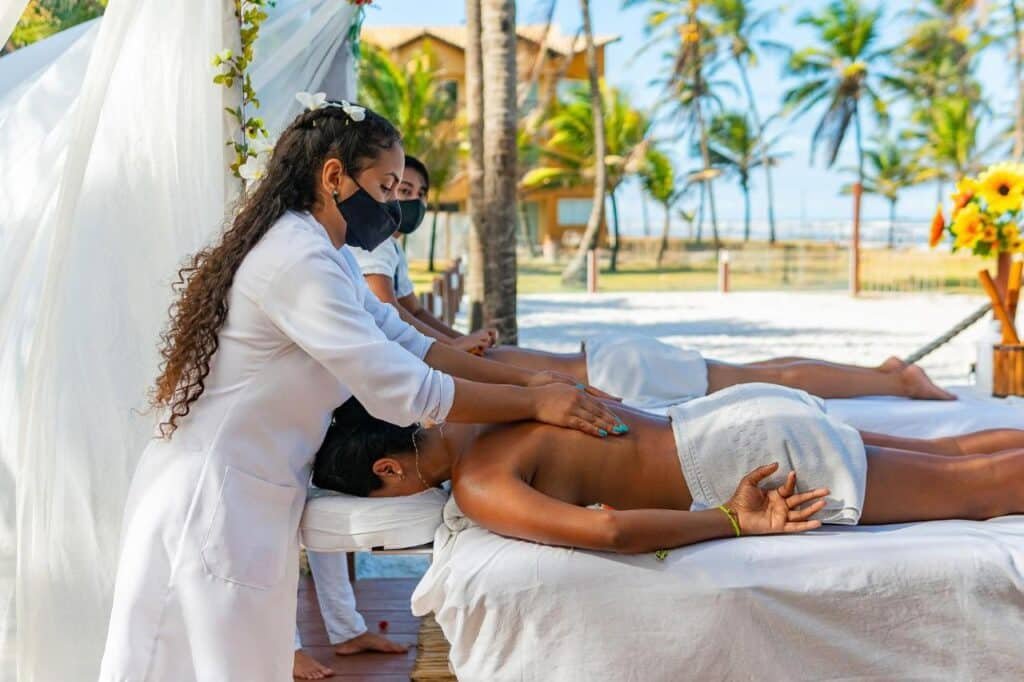 Duas pessoas recebendo massagens ao ar livre em um local à beira-mar no Makai Resort All Inclusive Convention Aracaju. Os massagistas usam máscaras e uniformes brancos.