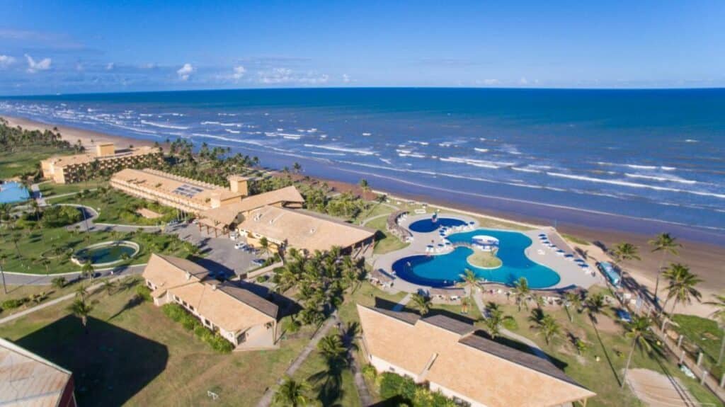 Vista aérea do Makai Resort All Inclusive Convention Aracaju com piscinas, cercado por palmeiras, perto de uma extensa praia de areia e oceano azul.