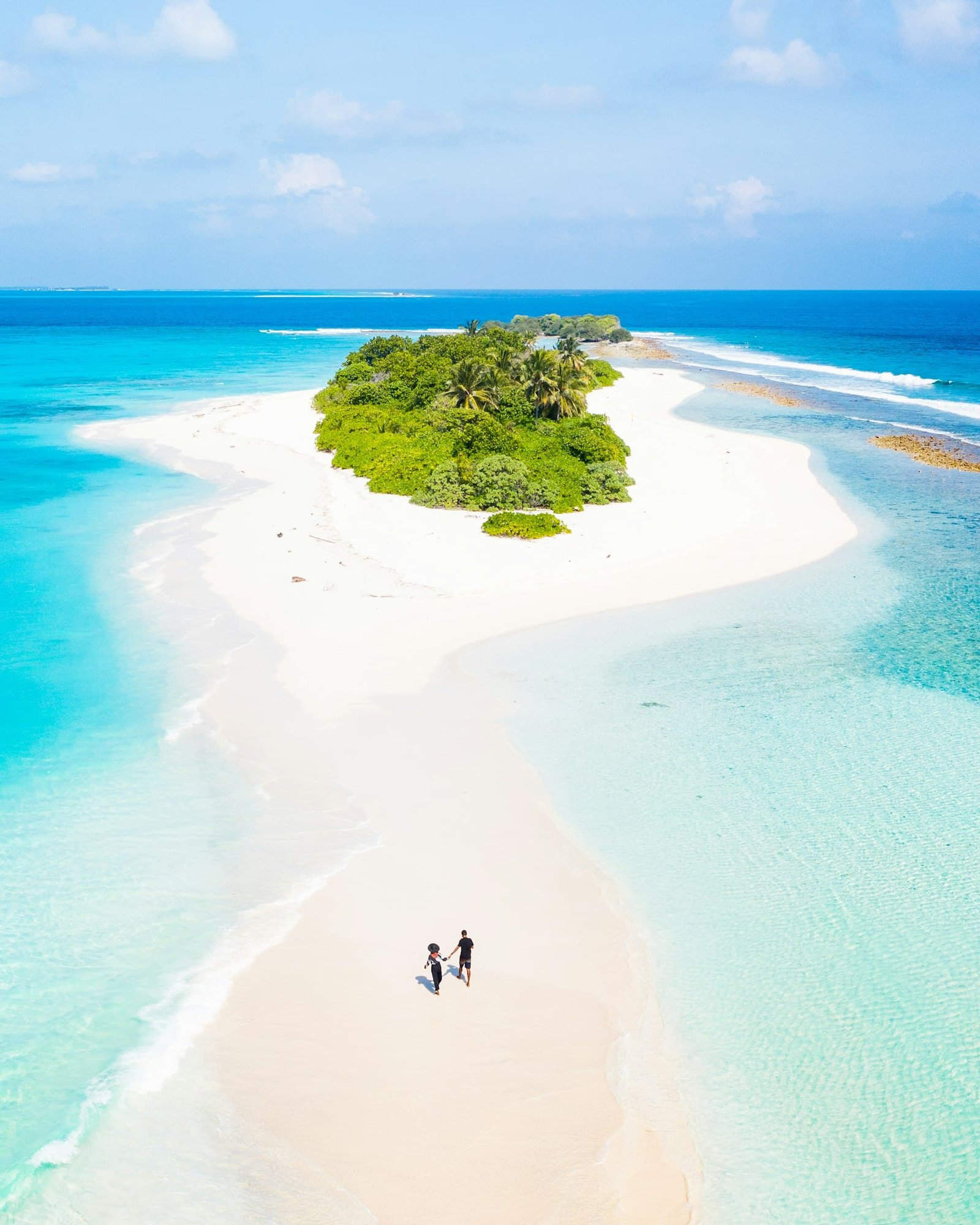 Vista aérea de um casal caminhando em uma praia de areia que leva a uma pequena e exuberante ilha cercada por águas azul-turquesa do oceano sob um céu azul.