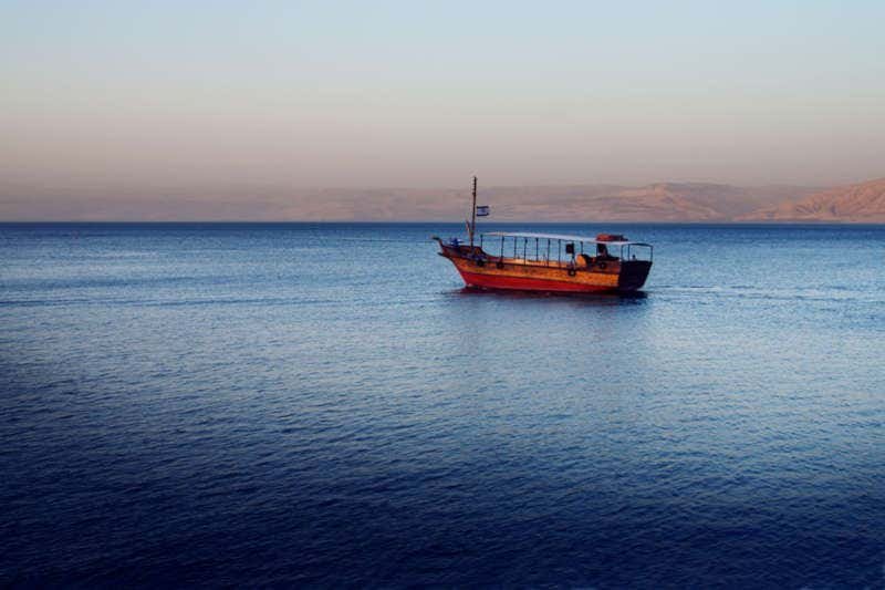 Um barco de madeira com cobertura navega em águas calmas e azuis do Mar da Galileia, com uma costa distante e colinas enevoadas sob um céu limpo. Representa o que fazer em Israel.