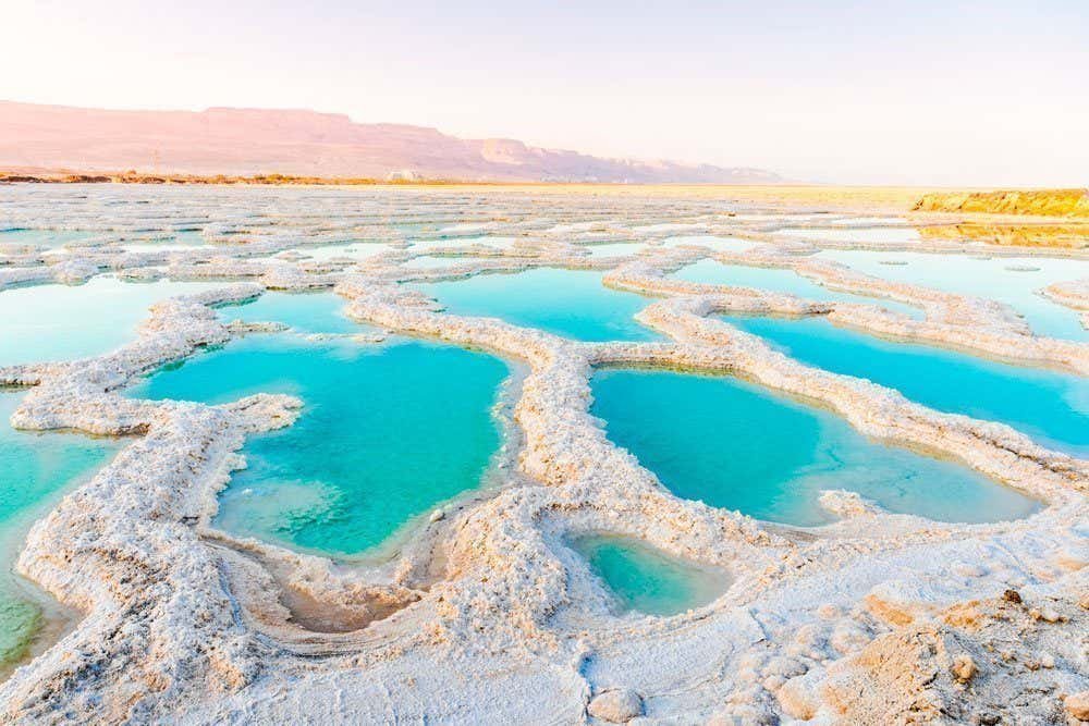 Formações de sal cercam piscinas de água azul-turquesa no Mar Morto em Israel, com colinas distantes sob um céu claro ao fundo.