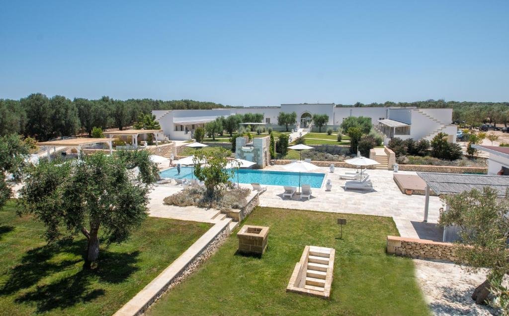 Foto aérea da Masseria Muntibianchi AgriResort que mostra algumas áreas de gramado com árvores verdes e outras parte com chão de piso e algumas construções. Há também uma piscina ao ar livre com algumas cadeiras e guarda-sóis durante o dia, ilustrando post Hotéis em Puglia.