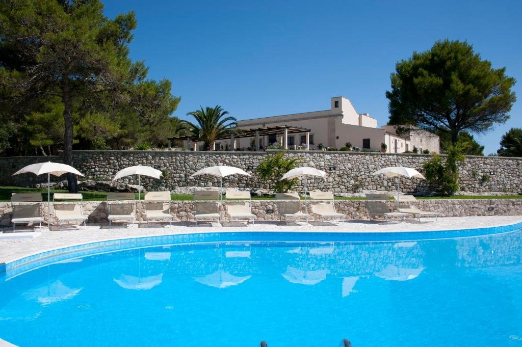 Parte da Masseria Panareo que mostra uma grande piscina azul com dez espreguiçadeiras e cinco guarda-sóis em volta. Além disso, tem uma grande parede de pedras atrás com algumas árvores verdes em volta e no fundo a construção da Masseria, ilustrando post Hotéis em Puglia. 