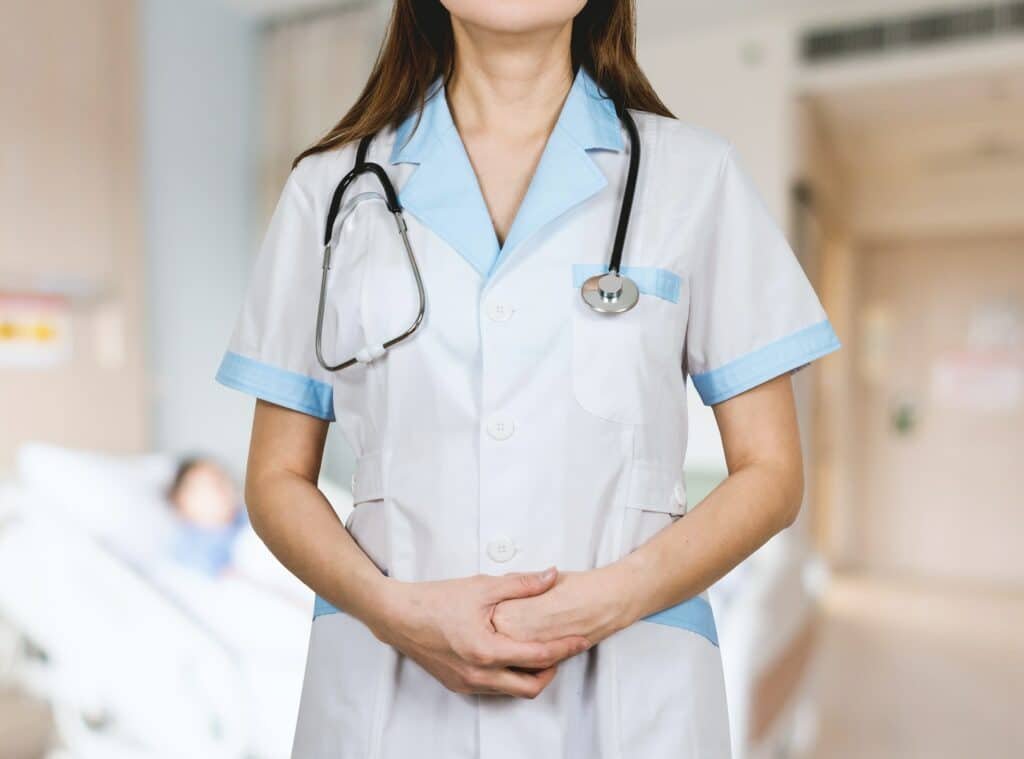 close em uma médica com estetoscópio no pescoço de mãos dadas e uniforme na frente de um leito com um paciente deitado desfocado