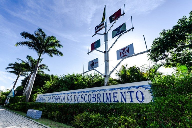 Imagem que mostra a frase "Memoria da Epopeia do Descobrimento" em uma placa em meio a arbustos. A foto representa o que fazer em Porto Seguro.