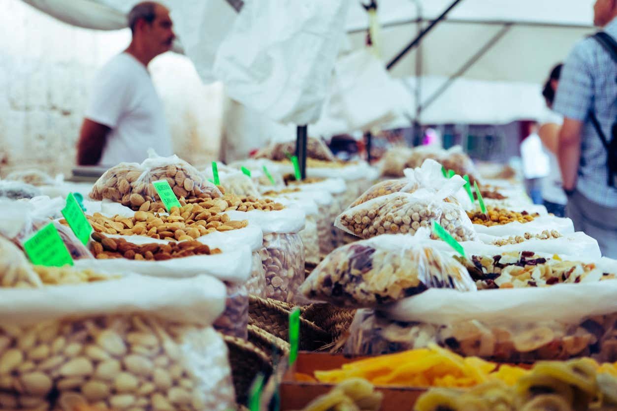 Sacos de nozes e frutas secas são exibidos em uma barraca de mercado sob um toldo. Um vendedor e um cliente são visíveis ao fundo.