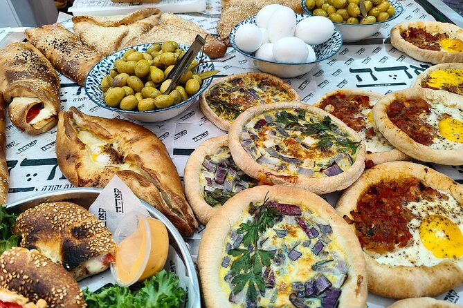 Uma variedade de produtos no Mercado Mahane Yehudd, sendo assados com coberturas, tigelas de azeitonas verdes e ovos cozidos em uma toalha de mesa estampada. Representa o que fazer em Israel.