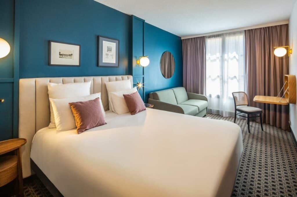 Quarto do Mercure Versailles Chateau, um dos hotéis perto do Palácio de Versalhes. Uma cama de casal com mesinhas de cabeceira e abajures dos dois lados está encostada na parede ao lado esquerdo. Ao lado direito da cama há um sofá com um espelho na parede acima que encara uma mesinha com cadeira. Há uma janela com cortinas na parede ao lado.