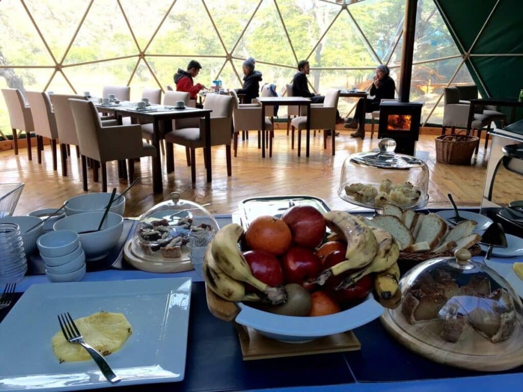 Mesa de café do Patagonia Eco Domesa frente com frutas, pães e bolos. Ao fundo cadeiras e mesas com pessoas.