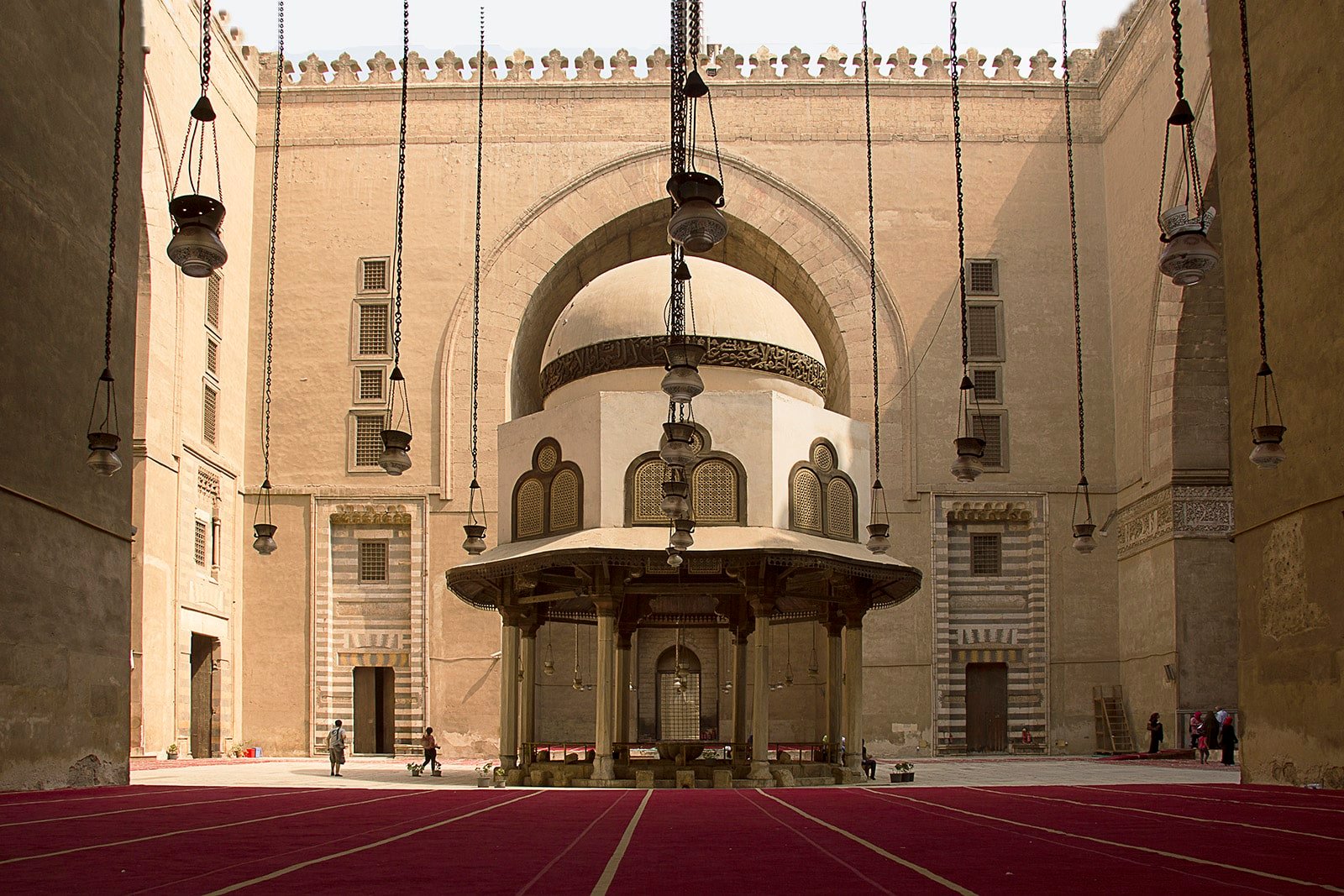 Pátio da Mesquita do Sultão Hassan com uma estrutura de cúpula central, cercada por muros e lanternas penduradas, com tapete vermelho cobrindo o chão. Imagem para ilustrar post sobre o que fazer no Cairo.