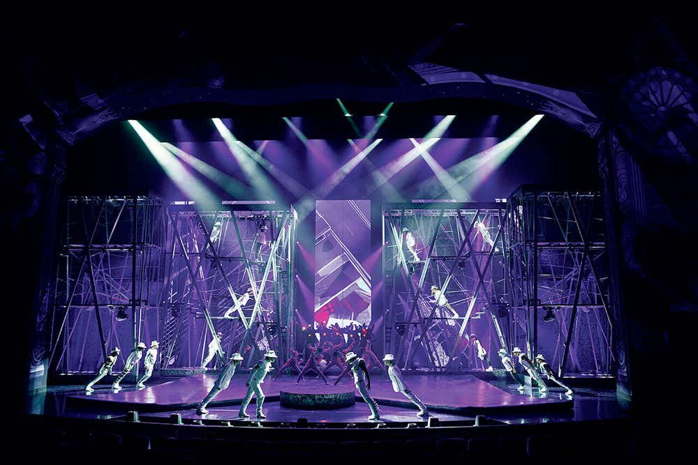 Performance de palco do Michael Jackson One com dançarinos em trajes brancos sob iluminação roxa. Cenário geométrico e andaimes em ambos os lados.