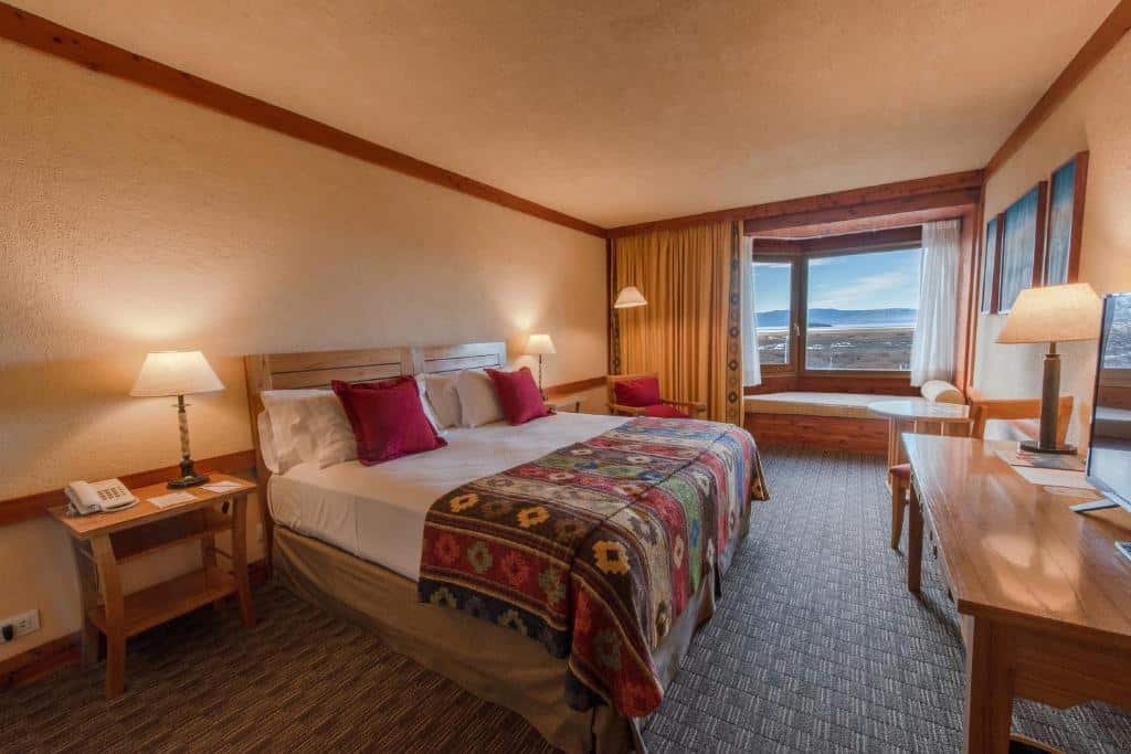 Quarto do Mirador del Lago Hotel. Uma cama de casal no lado esquerdo, com uma cômoda e um abajur de cada lado, de frente uma mesa redonda com uma cadeira e uma mesa com abajur e televisão. No fundo, no lado esquerdo, uma cadeira com um abajur, um sofá em baixo de uma janela com cortina.