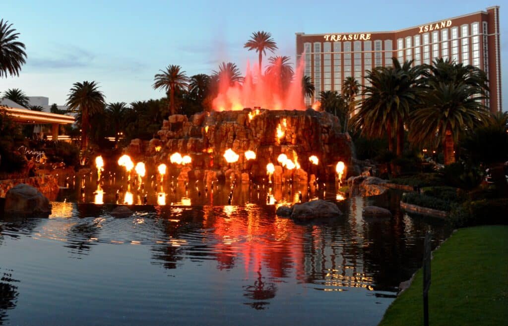 Foto do Mirage Volcano durante a erupção, cheio de chamas. Ele fica em meio a um lago e rodeado por um jardim. Esse é um dos pontos turísticos em Las Vegas.
