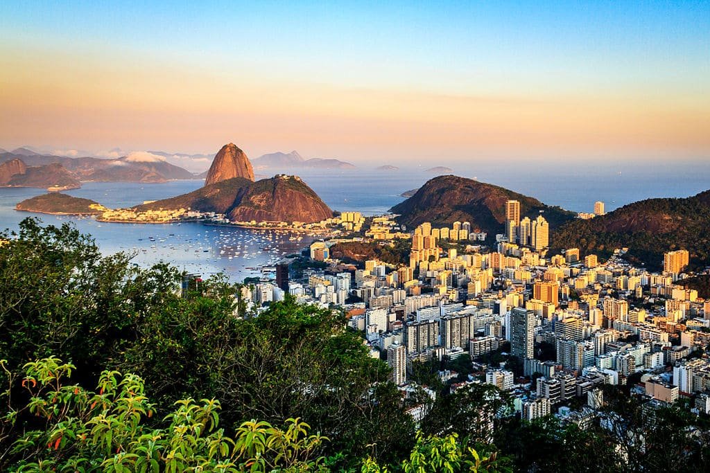 Vista aérea de uma paisagem urbana com uma baía, edifícios altos e uma montanha proeminente cercada por vegetação exuberante ao pôr do sol. Essa é a paisagem de uma das trilhas no Rio de Janeiro.