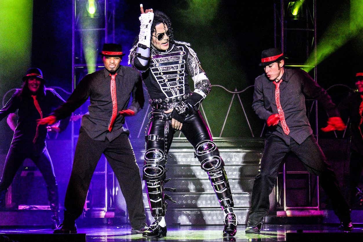 Sósia do Michael Jackson em um de seus trajes metálicos dança no palco com dois dançarinos de apoio em trajes pretos e detalhes em vermelho.