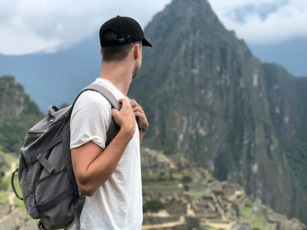 Homem de blusa branca e boné preto, olhando para a direita, com uma mochila nas costas e olhando para a montanha e cidade perdida de Machu Picchu. Representa o que levar na mala para o Peru