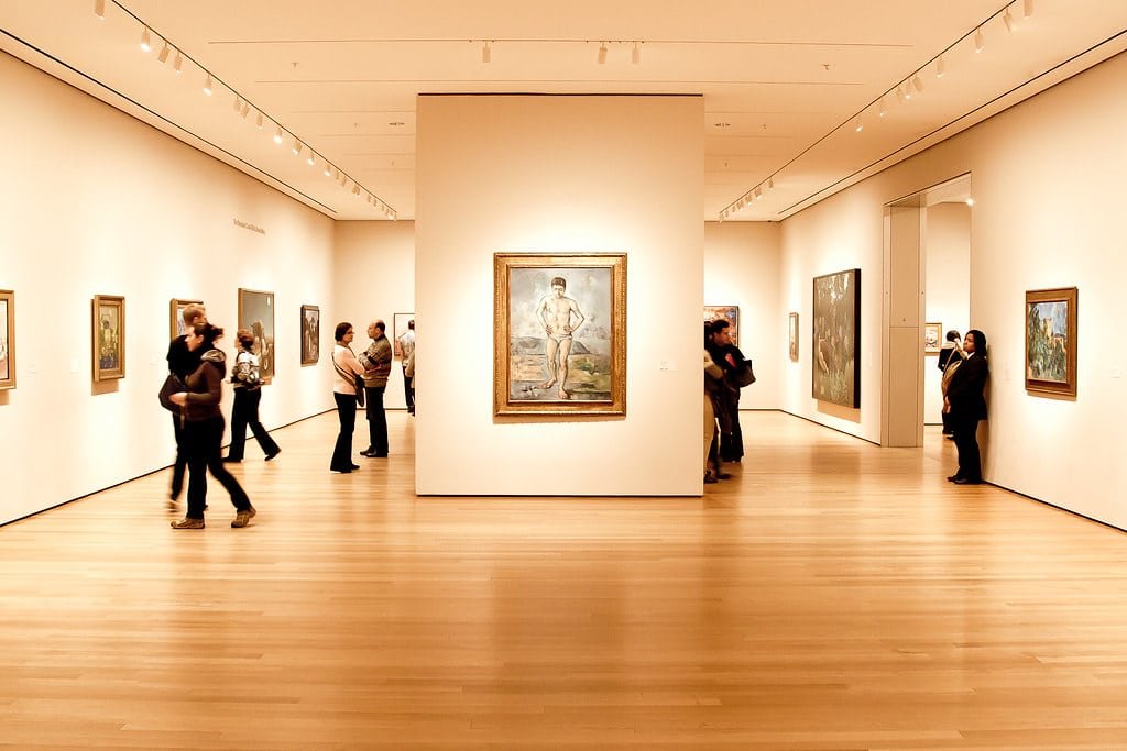Pessoas observando pinturas no MoMa - Museum of Modern Art com paredes brancas e pisos de madeira. Imagem para ilustrar post sobre museus em Nova York.