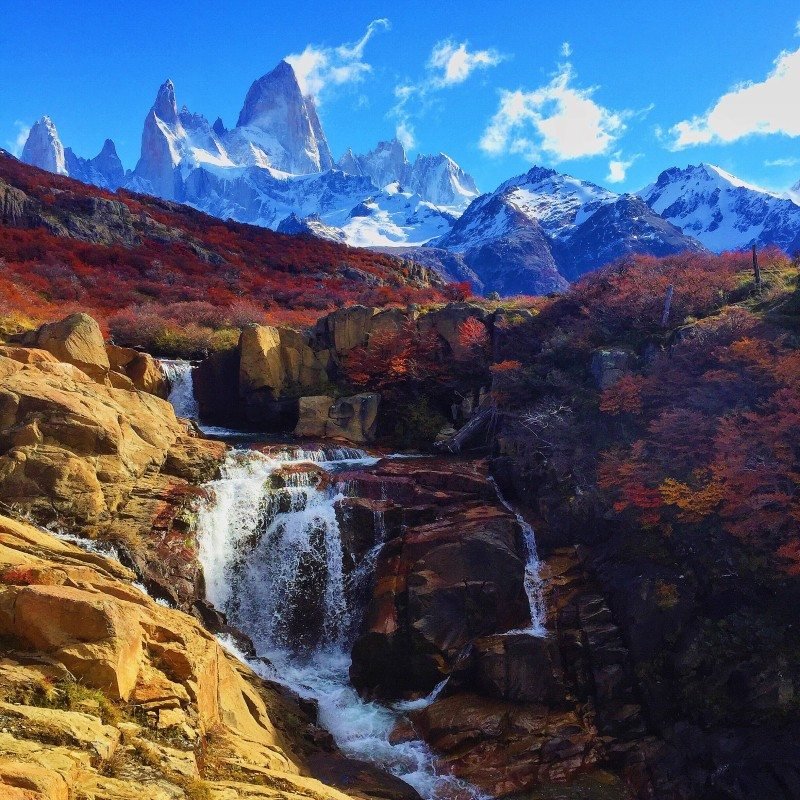 Uma cachoeira cai sobre rochas com montanhas cobertas de neve e árvores coloridas de outono ao fundo, sob um céu azul claro. Imagem para ilustrar post sobre a Patagônia Argentina.