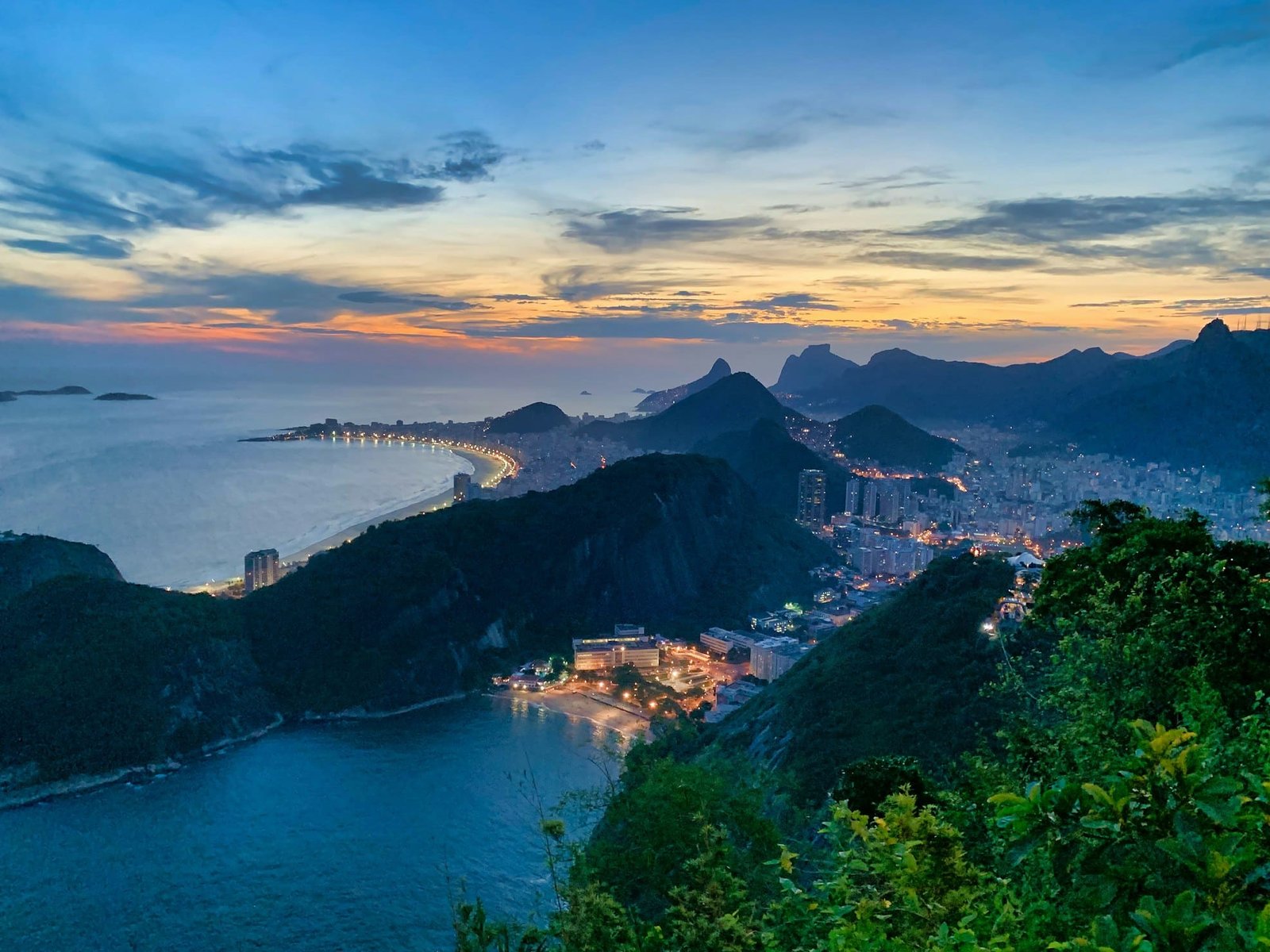 Vista aérea de uma cidade costeira ao anoitecer, com uma praia bem iluminada, colinas e um céu dramático com nuvens dispersas.