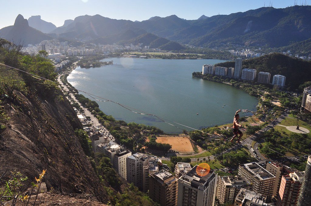 Pessoa fazendo tirolesa sobre uma paisagem urbana com um lago, montanhas e edifícios ao fundo, sob um céu limpo.