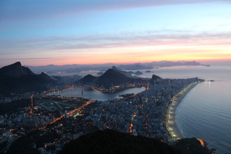 Vista aérea de uma paisagem urbana com litoral ao anoitecer, com ruas iluminadas, montanhas e um grande corpo d