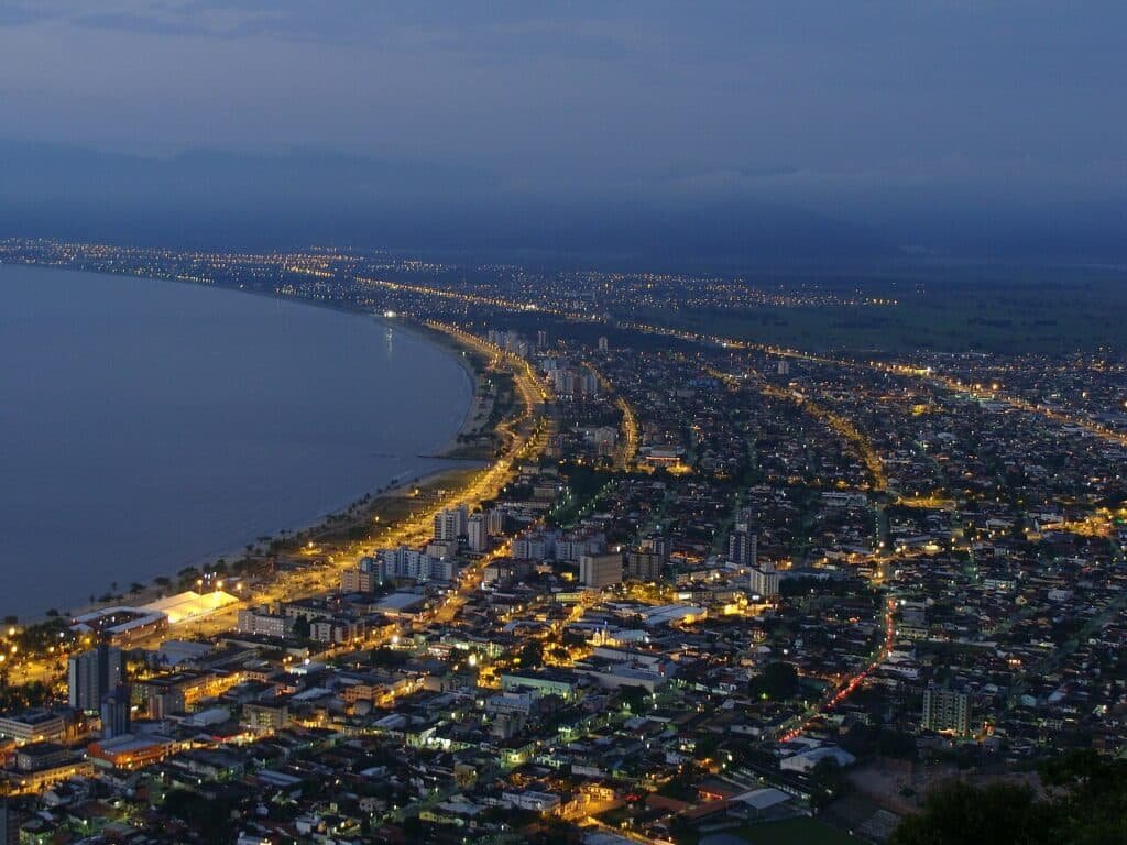 Vista aérea de uma cidade costeira ao anoitecer, com luzes da cidade iluminando ruas e edifícios ao longo da costa.