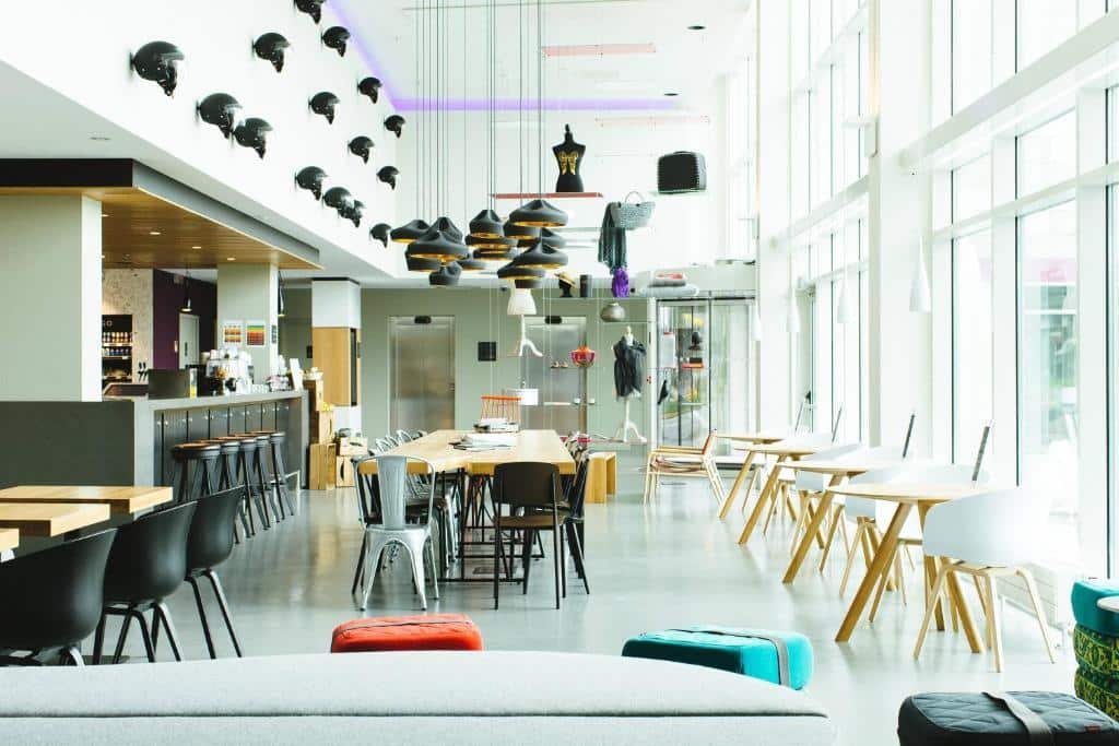 Salão de refeições do Moxy Milan Malpensa Airport com mesinhas redondas com cadeira, uma mesa extensa com cadeiras pretas, um balcão com banquinhos pretos e alguns itens de decoração 