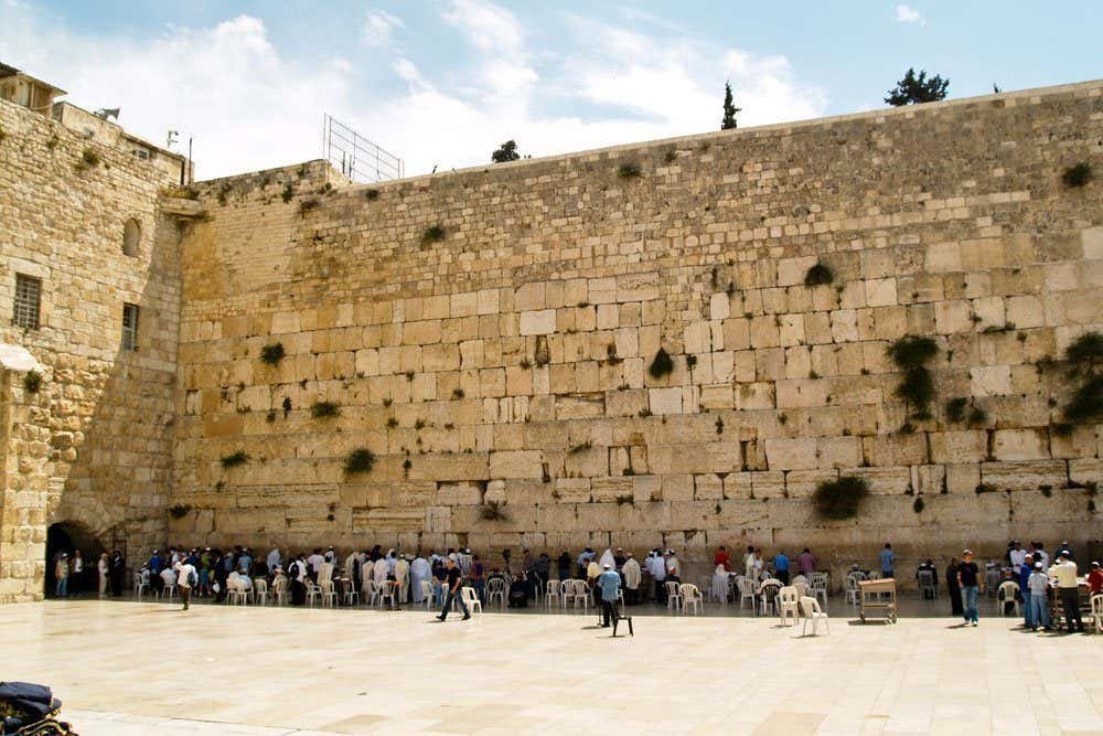 Pessoas se reúnem e rezam no Muro das Lamentações, uma antiga estrutura de calcário, sob um céu parcialmente nublado em Jerusalém.