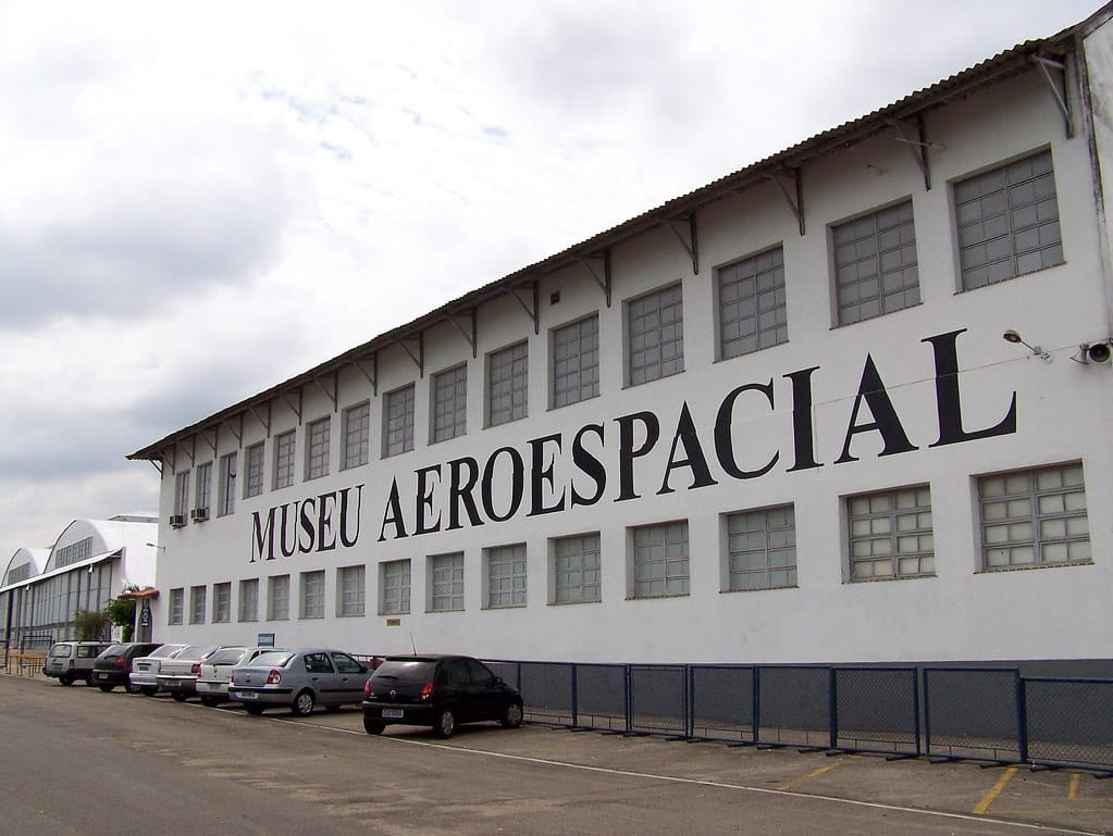 Vista externa do edifício do Museu Aeroespacial com estacionamento e vários carros estacionados. Imagem para ilustrar o post sobre museus no Rio de Janeiro.