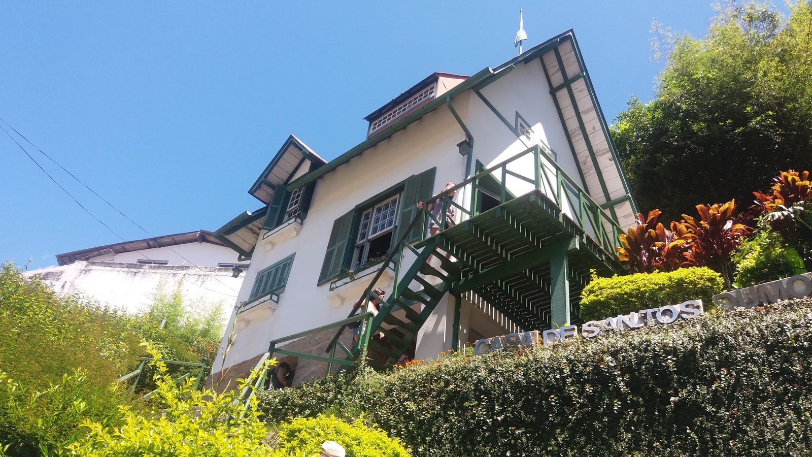 Uma casa branca com detalhes e escadas verdes é cercada por vegetação exuberante e arbustos, sob um céu azul claro.