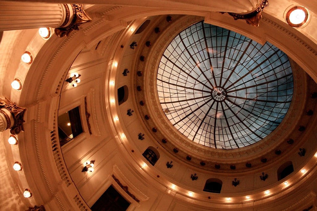 Interior do Centro Cultural Banco do Brasil com um grande teto de domo de vidro ornamentado, cercado por colunas e iluminado por luzes quentes.