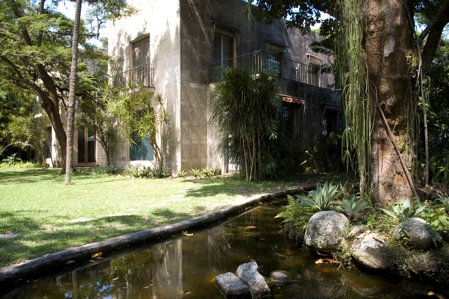 Um edifício de concreto cercado por árvores e plantas, com um pequeno lago em primeiro plano refletindo a vegetação. Imagem para ilustrar o post sobre museus no Rio de Janeiro.