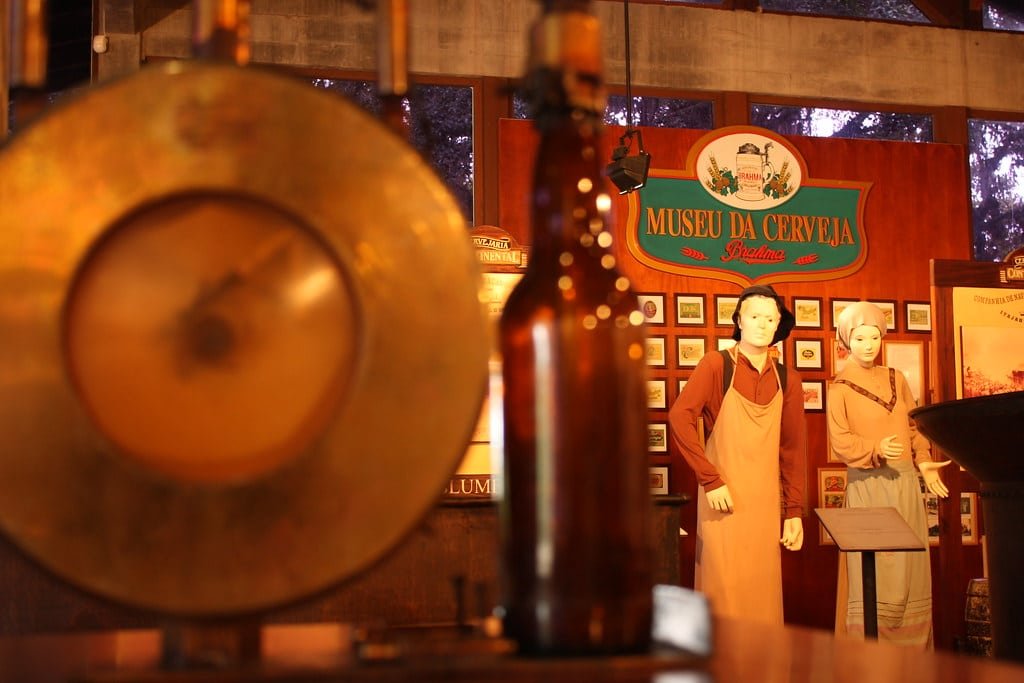 Exposição do Museu da Cerveja com manequins em trajes tradicionais, um grande gongo e uma garrafa de cerveja. Imagem para ilustrar post sobre o que fazer em Blumenau.
