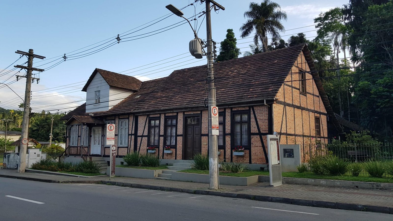 Uma casa tradicional com telhado de madeira e paredes de tijolos, situada ao lado de uma rua com postes de serviços públicos e cercada por vegetação.