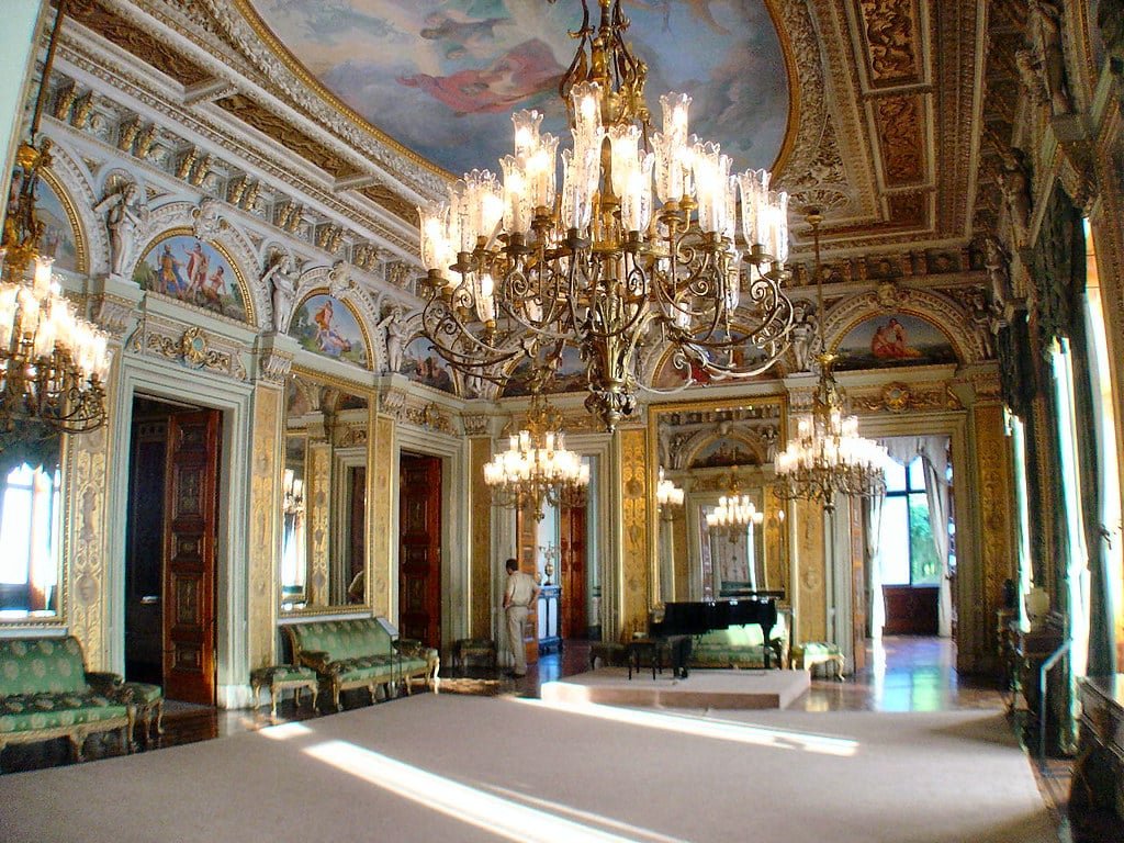 Sala ornamentada com lustres, um piano de cauda, pinturas decorativas e móveis opulentos, com janelas altas e tetos intrincados no Palácio do Catete. Imagem para ilustrar o post sobre museus no Rio de Janeiro.