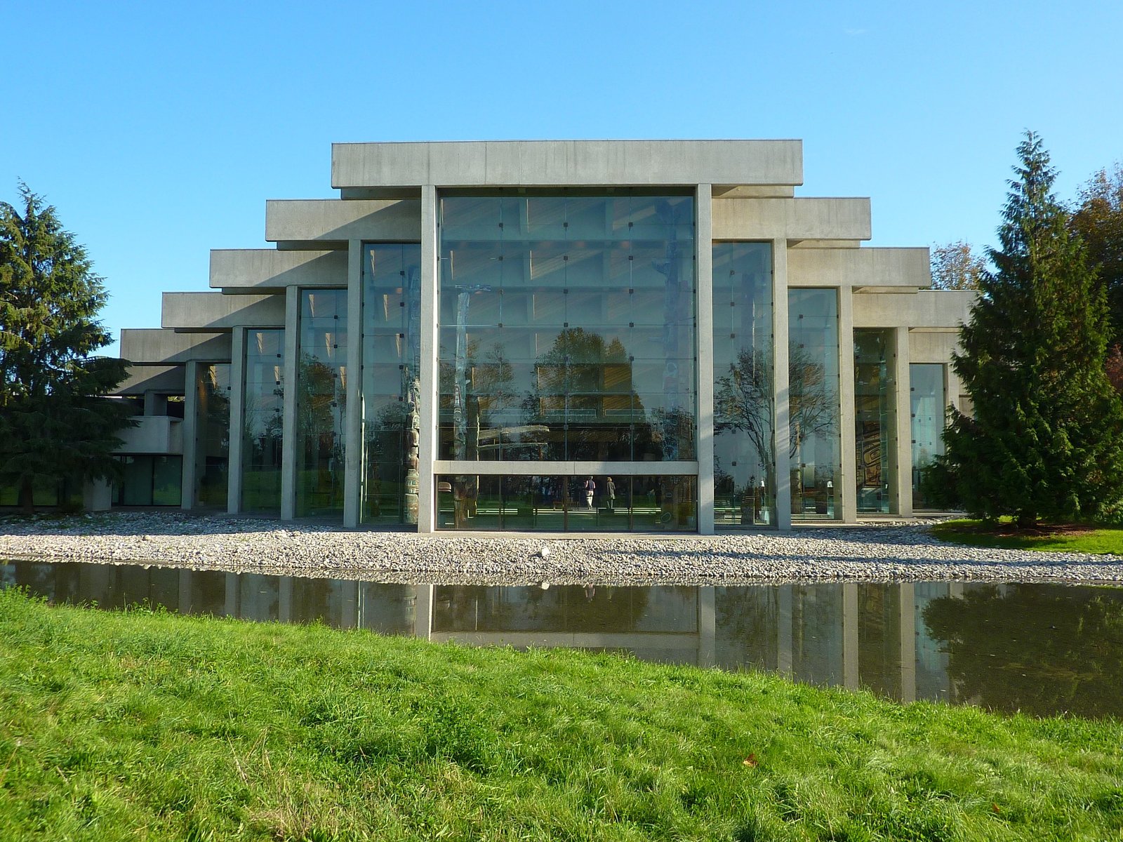 Museu de Antropologia (MOA) com grande fachada de vidro, tendo como pano de fundo árvores e um pequeno lago em primeiro plano em um dia ensolarado.