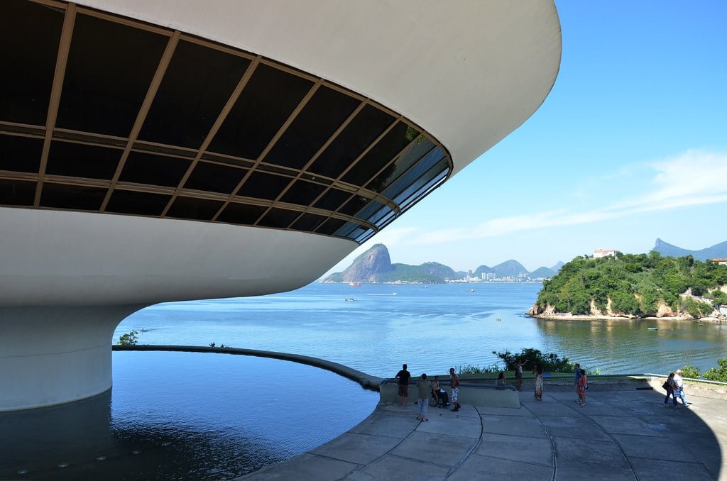 Edifício futurista do Museu de Arte Contemporânea com vista para a água e montanhas distantes, com pessoas caminhando por perto sob um céu azul claro. Imagem para ilustrar o post sobre museus no Rio de Janeiro.