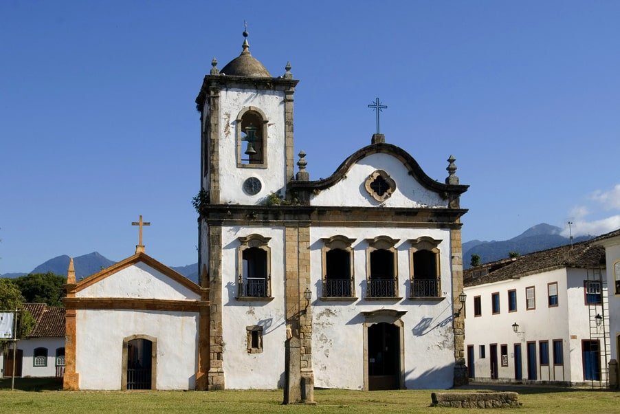 A imagem mostra a Igreja de Santa Rita de Cássia, um dos marcos históricos de Paraty. A igreja possui uma fachada colonial simples, com paredes brancas e detalhes em pedra. A estrutura principal inclui uma torre sineira à esquerda e uma entrada central com uma pequena porta e janelas acima. Há um cruzeiro de pedra em frente à igreja. O céu está claro, destacando ainda mais a beleza arquitetônica da construção. Esta igreja é um importante ponto turístico e histórico da cidade de Paraty.