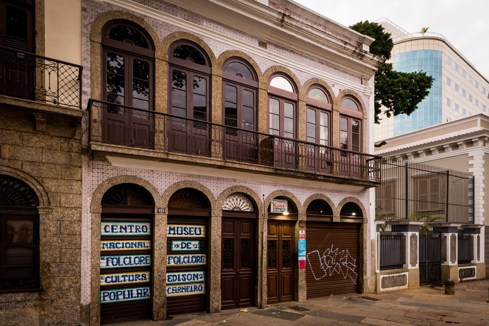 Prédio histórico com janelas arqueadas e grafite, com placas de um museu de folclore. Prédio moderno visível ao fundo. Imagem para ilustrar o post sobre museus no Rio de Janeiro.