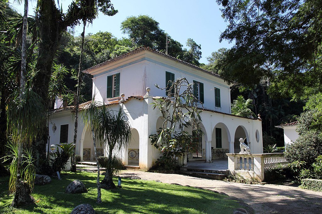 Casa branca em estilo colonial com persianas verdes, cercada por exuberante vegetação tropical, incluindo grandes árvores e diversas plantas.