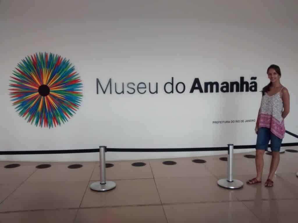 Uma mulher está ao lado da placa "Museu do Amanhã" em uma parede branca, com um desenho circular colorido à esquerda do texto.