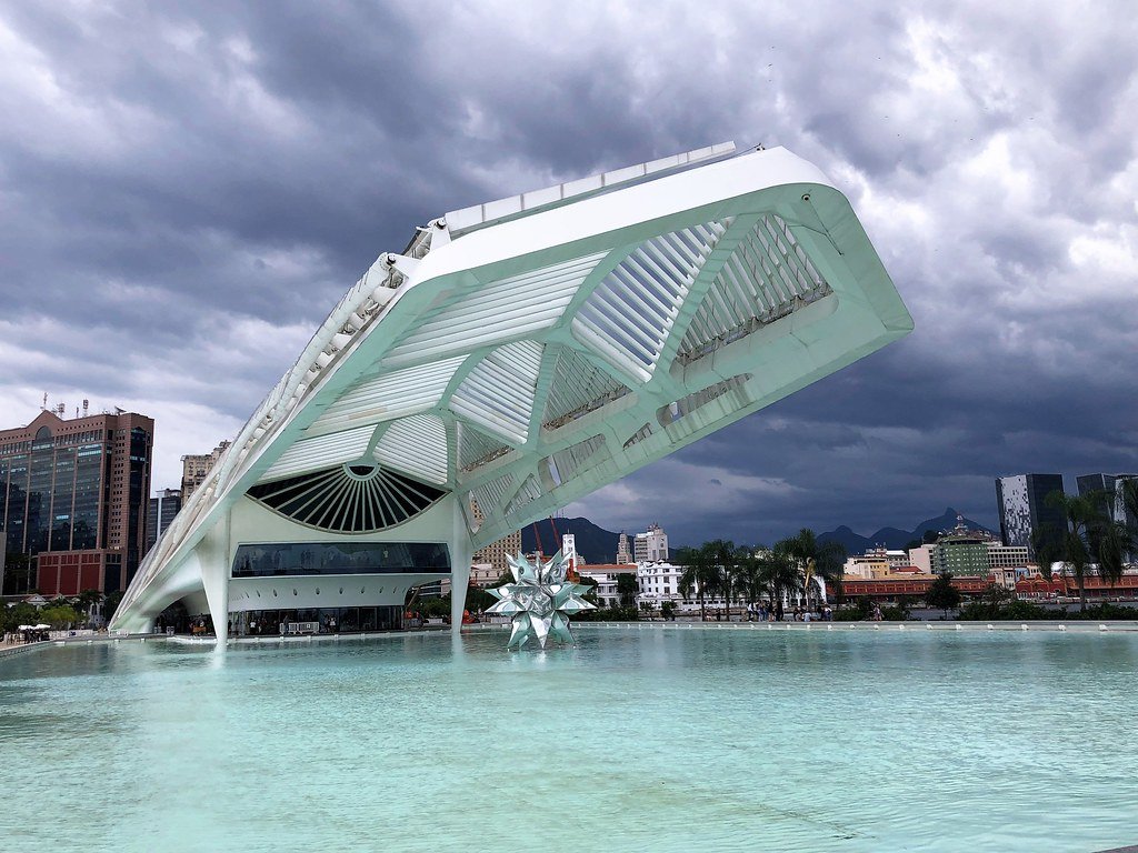 Museu do Amanhã com um desenho geométrico perto de uma piscina de água, com céu nublado e prédios urbanos ao fundo. Imagem para ilustrar o post sobre museus no Rio de Janeiro.