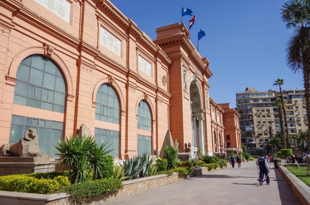 Exterior do Museu Egípicio rosa com janelas em arco, bandeiras no topo e pessoas caminhando pelo caminho ladeado por vegetação. Imagem para ilustrar post sobre o que fazer no Cairo.