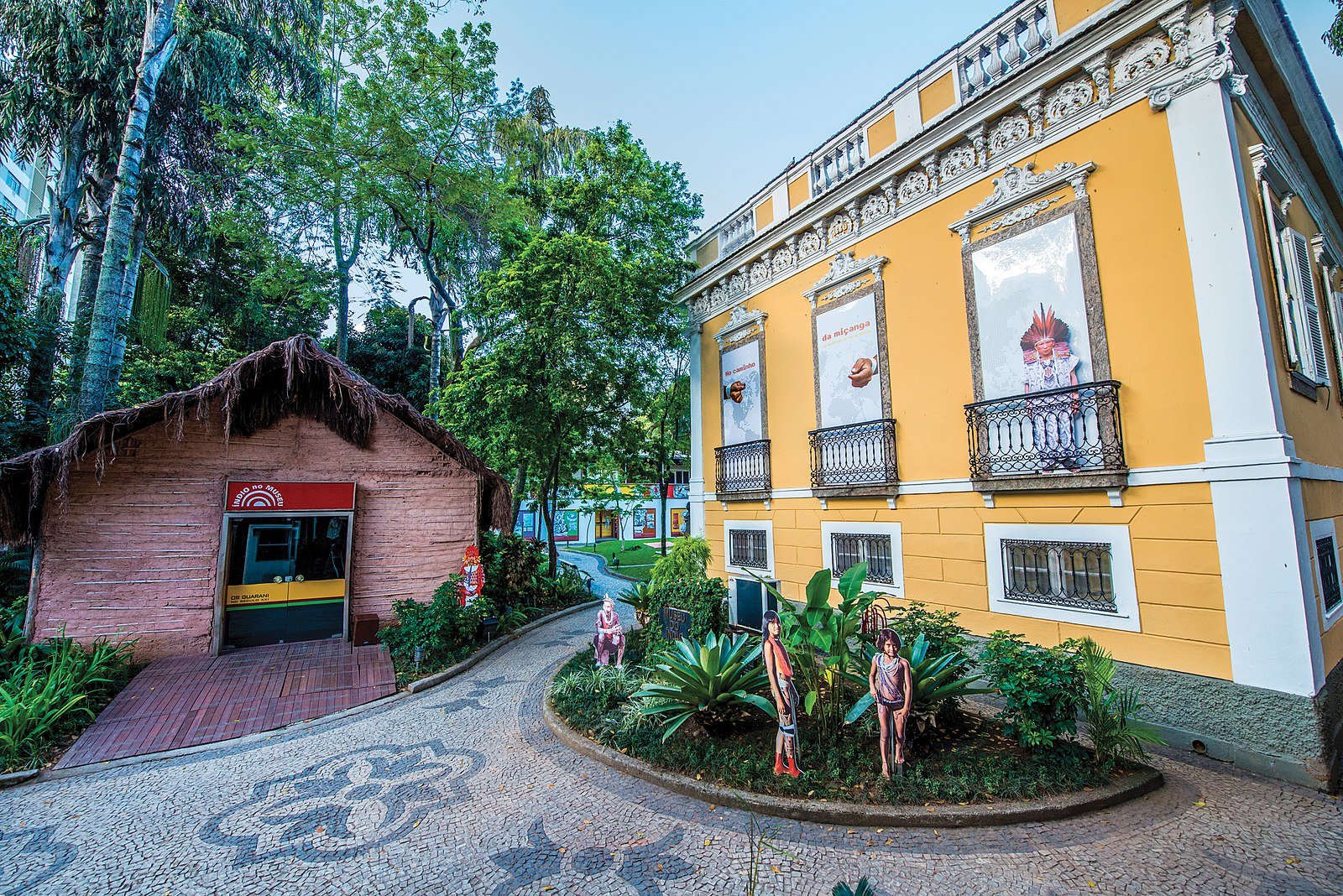 Um prédio amarelo com detalhes ornamentados fica ao lado de uma cabana de palha. Manequins e plantas decoram o caminho de azulejos no meio. Imagem para ilustrar o post sobre museus no Rio de Janeiro.