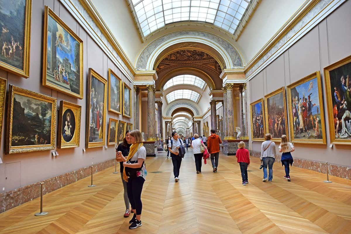 Um corredor do museu do Louvre com pinturas em ambos os lados. As pessoas andam e observam as obras de arte. O teto apresenta grandes claraboias arqueadas.